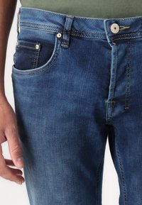 Tummansiniset farkut, joissa on klassinen viiden taskun malli, näkyvä ommelkuvio ja metallinen nappisuljenta vyötäröllä.