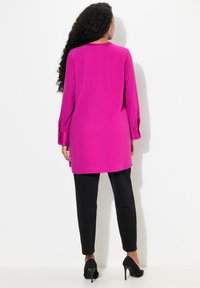 Helle magenta Tunika mit langen Ärmeln, abgerundetem Saum und geknöpften Manschetten, kombiniert mit schwarz, eng anliegenden Hosen und schwarzen High Heels, von hinten gezeigt.