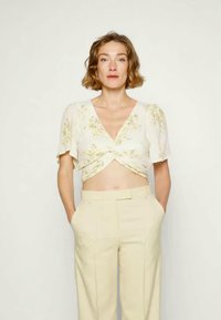 Top cropped com padrão floral em creme, com mangas curtas em puff e frente torcida, combinado com calças de cintura alta em beige claro.