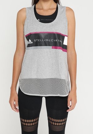 Woman wearing sleeveless gray Adidas Stella McCartney sports top with mesh hem and black leggings with patterned cutouts.