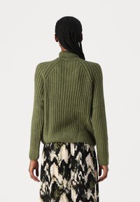 Suéter acanalado de color verde oliva con un alto cuello y mangas largas, que presenta un ajuste ceñido. Llevado sobre una falda con un patrón en blanco y negro.