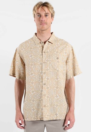 Homme blond portant une chemise beige à manches courtes avec un motif circulaire blanc complexe et une poche poitrine.