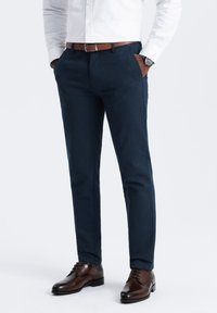 Pantaloni slim-fit blu navy realizzati in tessuto strutturato, con tasche laterali e cintura in pelle marrone; abbinati a scarpe eleganti marroni.