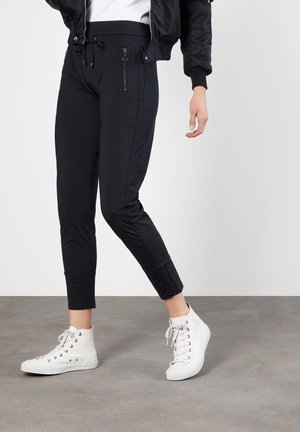 Person, die schwarze Jogginghosen mit Reißverschlusstaschen und weiße High-Top-Sneaker trägt, steht auf grauem Boden vor weißem Hintergrund.