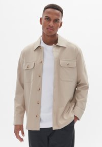 Camisa de botones beige en tejido suave con dos bolsillos en el pecho, botones marrones y un cuello, llevada sobre una camiseta blanca, combinada con pantalones oscuros.