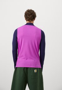 Højhalset ærmeløs top med en tekstureret pink front og blå lange ærmer, parret med grønne bukser med et logo-patch.