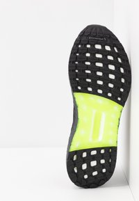 Czarna podeszwa butów biegowych w kolorze neonowej żółci z bieżnikiem Continental, pochylona pionowo wzdłuż białej ściany w rogu.