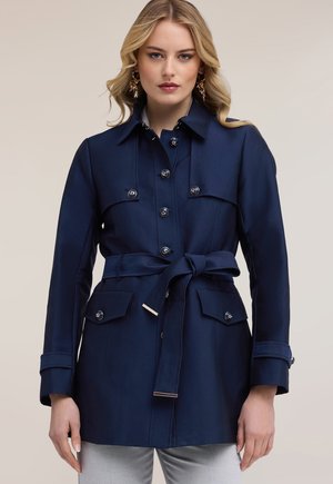 Mujer rubia con gabardina azul marino ceñida con cinturón y detalles de botones, lleva pendientes dorados en forma de hoja, sobre un fondo neutro.