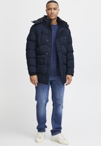 Navy blue puffer jacket with a hood, featuring front pockets and a zip closure, layered over a dark blue sweatshirt and worn with blue jeans.