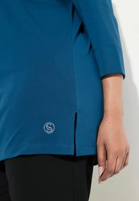 Blå tunika med 3/4 ärmar, tillverkad av mjukt tyg. Har en sidoklyfta och en cirkulär logo prydd med rhinstenar vid fållen.