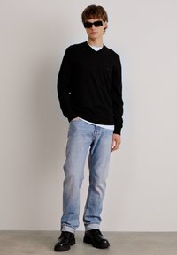 Schwarzer V-Ausschnitt-Pullover, hellblaue Jeans und schwarze Schuhe. Sonnenbrillen verleihen einen modernen Touch. Einfaches Design mit minimalen Details.