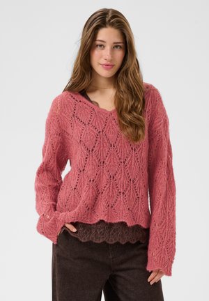 Young woman with long brown hair wearing a pink patterned knit sweater and black pants, standing with one hand in pocket.