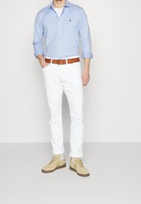 Vaaleansininen pitkähihaisen nappipaidan, valkoiset farkut, ruskea vyö ja beige urheilukengät, joissa on ruskeita yksityiskohtia, seisoo harmaata taustaa vasten.