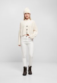 Vit chunky stickad cardigan med svarta knappar över en vit mock neck-top, kombinerat med vita skinny jeans och bruna snörskor.