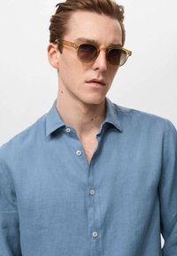 Light blue linen shirt with a relaxed collar, buttoned front, and subtle texture; paired with brown-tinted sunglasses with clear frames.
