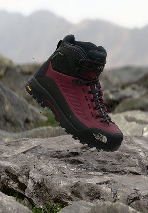 Bota de senderismo The North Face en burdeos y negro en el aire sobre terreno rocoso con fondo de montaña desenfocado.