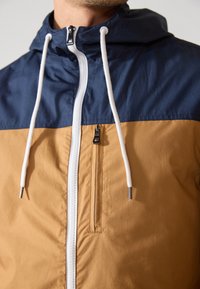Farvet blazer med en mørkeblå øverste del og en brun nederste del. Har en frontlynlås, justerbar hætte og to lynlåsede lommer.