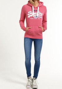 Sudadera rosa con capucha, que presenta un logo blanco de "SuperDry" y bolsillos delanteros. Combinada con jeans ajustados azules y zapatillas blancas.
