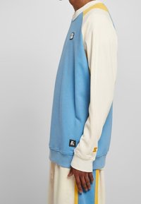 Uomo che indossa una felpa a maniche lunghe blu e crema con accenti gialli, in piedi di lato con la mano rilassata lungo il corpo.