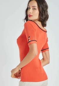 Suéter ajustado de canalé en vibrante color coral con un escote amplio, mangas cortas, ribetes negros y botones decorativos en los puños.