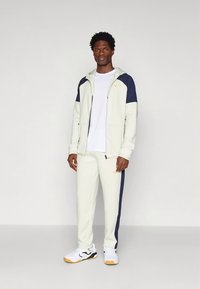 Svetlosivá tepláková súprava s námorníckymi akcentmi, zahŕňajúca bundu so zipsom a zúžené nohavice. Vyrobená z hladkej látky, so logom na hrudi a boku.