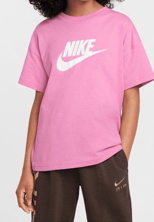 Kind trägt ein pinkes Nike T-Shirt mit weißem Logo und dunkelbraune Nike Air Hose mit Logo am linken Oberschenkel, steht mit einer Hand in der Tasche.