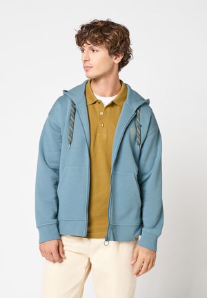 Homme aux cheveux bouclés portant un sweat à capuche bleu zippé par-dessus un polo moutarde et un pantalon beige, regardant vers sa gauche.