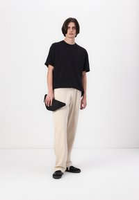 Zwarte katoenen t-shirt met een zak, gecombineerd met losse beige broek. Houdt een zwarte ritssluiting tas vast. Zwarte schoenen maken de outfit af.