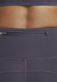 Leggings de sport violet foncé avec une taille plate, dotés d'une poche zippée à l'arrière et de coutures visibles.