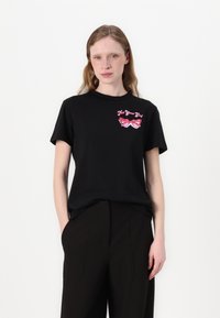 Musta puuvillainen t-paita lyhyillä hihoilla, pyöreällä kaula-aukolla ja etupuolella graafinen kuva vaaleanpunaisista aurinkolasista ja teksti "Not Your Girl". Malli seisoo kädet taskuissa.
