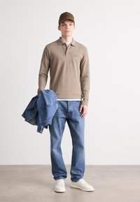 Camisa polo de mangas longas bege, calças de ganga azuis, sapatilhas brancas e um boné combinando segurado com uma jaqueta de ganga ao braço.