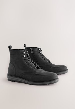 Cizme scurte negre din piele întoarsă cu textură fină, șireturi în față și fermoar lateral. Au talpă de cauciuc și detalii cusute pe vârf.