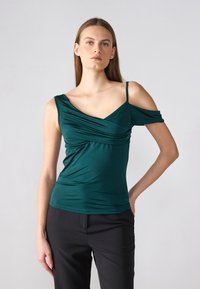 Tyrkysový, přiléhavý top s drapovanými detaily, off-shoulder rukávy a tenkými ramínky. V kombinaci s černými kalhotami. Hladká textura.
