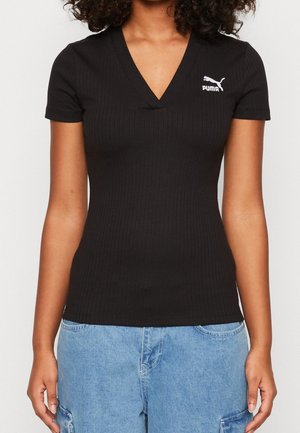 Kobieta nosząca dopasowany czarny prążkowany T-shirt Puma z dekoltem w serek w połączeniu z jasnoniebieskimi denimowymi spodniami cargo, ramiona swobodnie opuszczone wzdłuż ciała.
