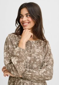 Blumenmusterkleid in Hellbraun mit cremefarbenen Blumenmustern. Es hat einen Bootsausschnitt und elastische Bündchen an den langen, puffartigen Ärmeln.