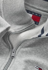 Sudadera ligera de color gris claro con cierre de cremallera, cuello acanalado, forro de felpa y logotipo de Tommy Hilfiger en el pecho y etiqueta en el interior del cuello.