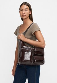 Frau in taupefarbenem Kurzarmoberteil und dunklen Jeans, die eine gro�ße dunkelbraune Lederumhängetasche mit mehreren vorderen Taschen hält.