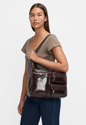 Donna che indossa un top a maniche corte color taupe e jeans scuri, mentre tiene una grande borsa a spalla in pelle marrone scuro con più tasche frontali.