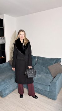 Casaco preto com gola de pele sintética, design ajustado. Calças largas burgundy, bolsa preta e sapatos envernizados. Sofá de tecido suave em tom verde-água ao fundo.