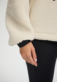 Cremfarbener Fleecejacke mit strukturierter Oberfläche, weiten Ärmeln und einem schwarzen Bündchen, kombiniert mit eleganten schwarzen Leggings.