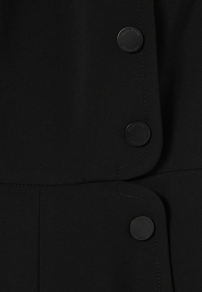 Vêtement noir en tissu lisse, avec des bords arrondis et de boutons noirs ronds portant un texte en relief. Les détails de couture sont visibles le long des coutures.