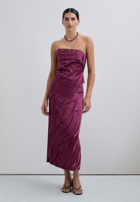 Vestido sin tirantes, de largo hasta el suelo, en satén magenta intenso con un patrón texturizado. Presenta un corsé ajustado y una falda fluida. Tacones negros con tiras.