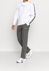 Uomo che indossa un sudore bianco Champion con strisce logo sulle maniche, pantaloni della tuta grigi Champion e sneaker bianche Nike, in piedi su uno sfondo neutro.