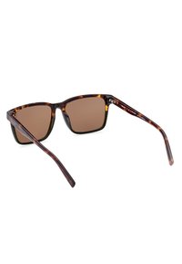 Occhiali da sole con tonalità marrone e montatura a tartaruga. Lenti quadrate e aste sottili, con una finitura lucida e dettagli hardware minimali.