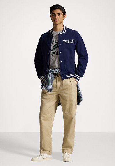 Blauwe varsity jacket met witte gestreepte manchetten, grijs T-shirt met logo, kaki broek en een geruite blouse om de taille geknoopt. Crème sneakers.