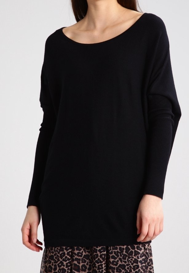 Femme portant un haut noir à manches longues ample avec un col large et une jupe à imprimé léopard sur un fond uni.