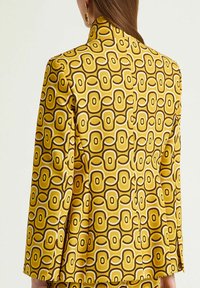 Blazer à motifs jaunes avec un design rétro et des accents marron et blancs. Le tissu a une texture lisse et un col structuré.