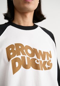 Weißes T-Shirt mit schwarzen Raglanärmeln, versehen mit kräftigem braunem Text "BROWN DUCKS." Glatte Baumwollstruktur. Nahaufnahme des Halsbereichs.