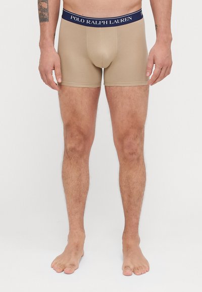 Bež bokserice s elastičnim mornarsko modrim pasom označenim "Polo Ralph Lauren". Gladka tekstura, prilega se telesu in reza se do sredine stegna.