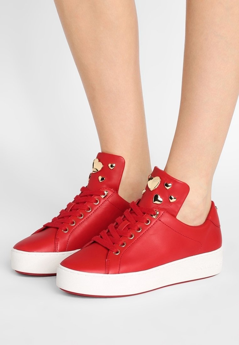 Baskets en cuir rouge avec semelles épaisses blanches, clous dorés en forme de cœur sur la languette, portées pieds nus sur un fond uni.